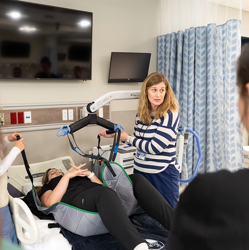 A faculty member demonstrates the use of a patient lift in a healthcare training room. A student is seated in the lift while others observe the demonstration.