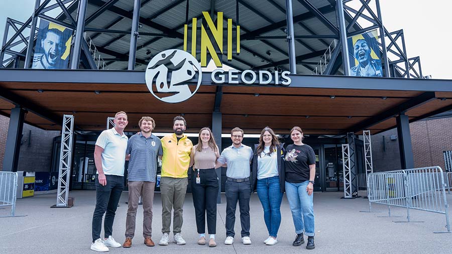 A group of Belmont University graduates stands in front of the Geodis Park entrance, home of Nashville SC, with the club's iconic yellow logo prominently displayed above them.