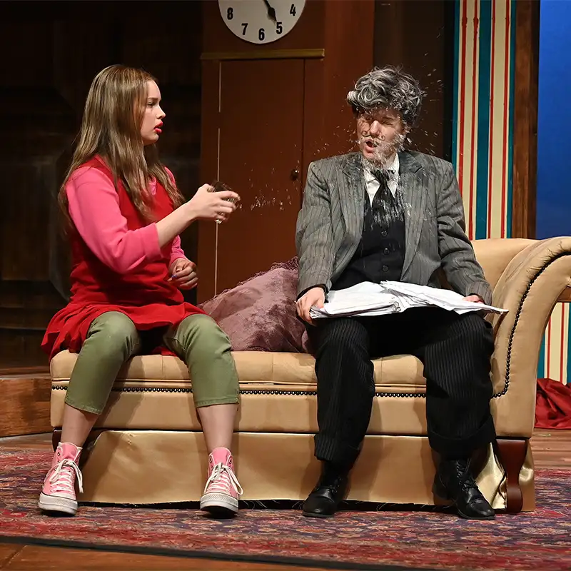 A scene from a play featuring a young woman in a red sweater and green pants sitting on a couch, holding a small object, while an older man in a suit with a messy hairstyle reacts with surprise as something splashes onto him. A clock is visible in the background.