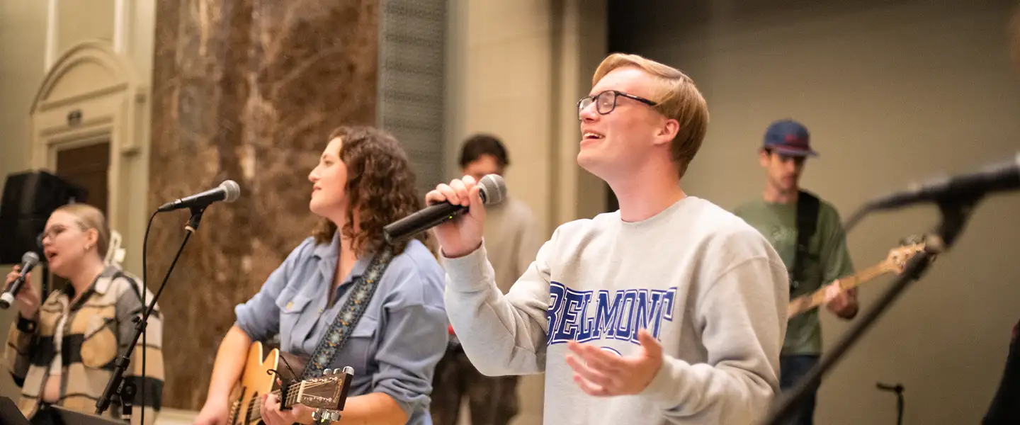 A group of musicians performing on stage, with one male singer passionately singing into a microphone while wearing a Belmont sweatshirt, and a female guitarist beside him. Other band members are visible in the background.