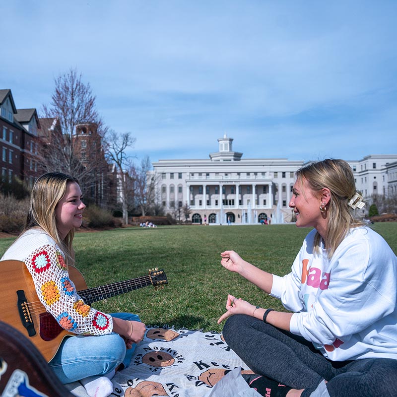 2 students sitting on the lawn while collaboration over song as one of them plays the guitar