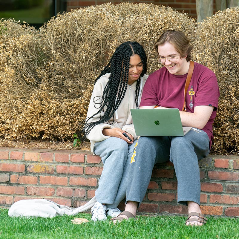 2 students sit together and look at laptop outside