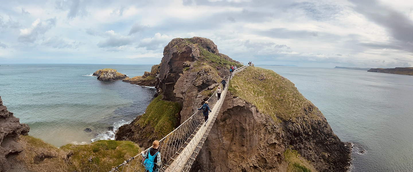 People walk across a narrow rope bridge spanning rugged sea cliffs along the coast of Ireland, with rocky islands, green grass, and the ocean stretching out beneath a cloudy sky.