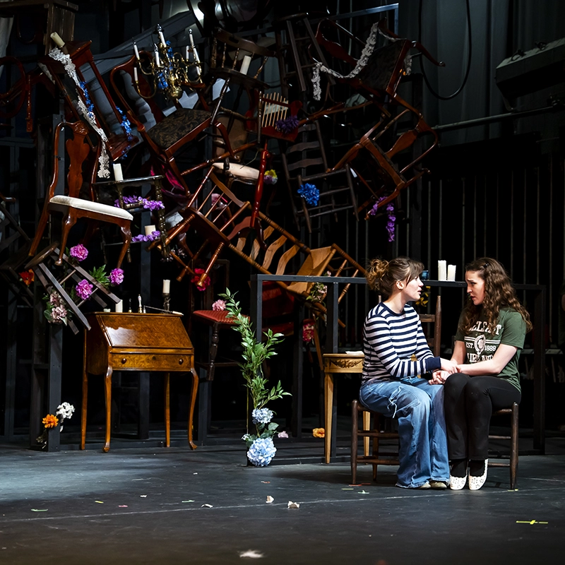wo women sit on chairs on a theater stage, holding hands, and acting out a scene amidst a dramatic backdrop of overturned chairs and furniture.