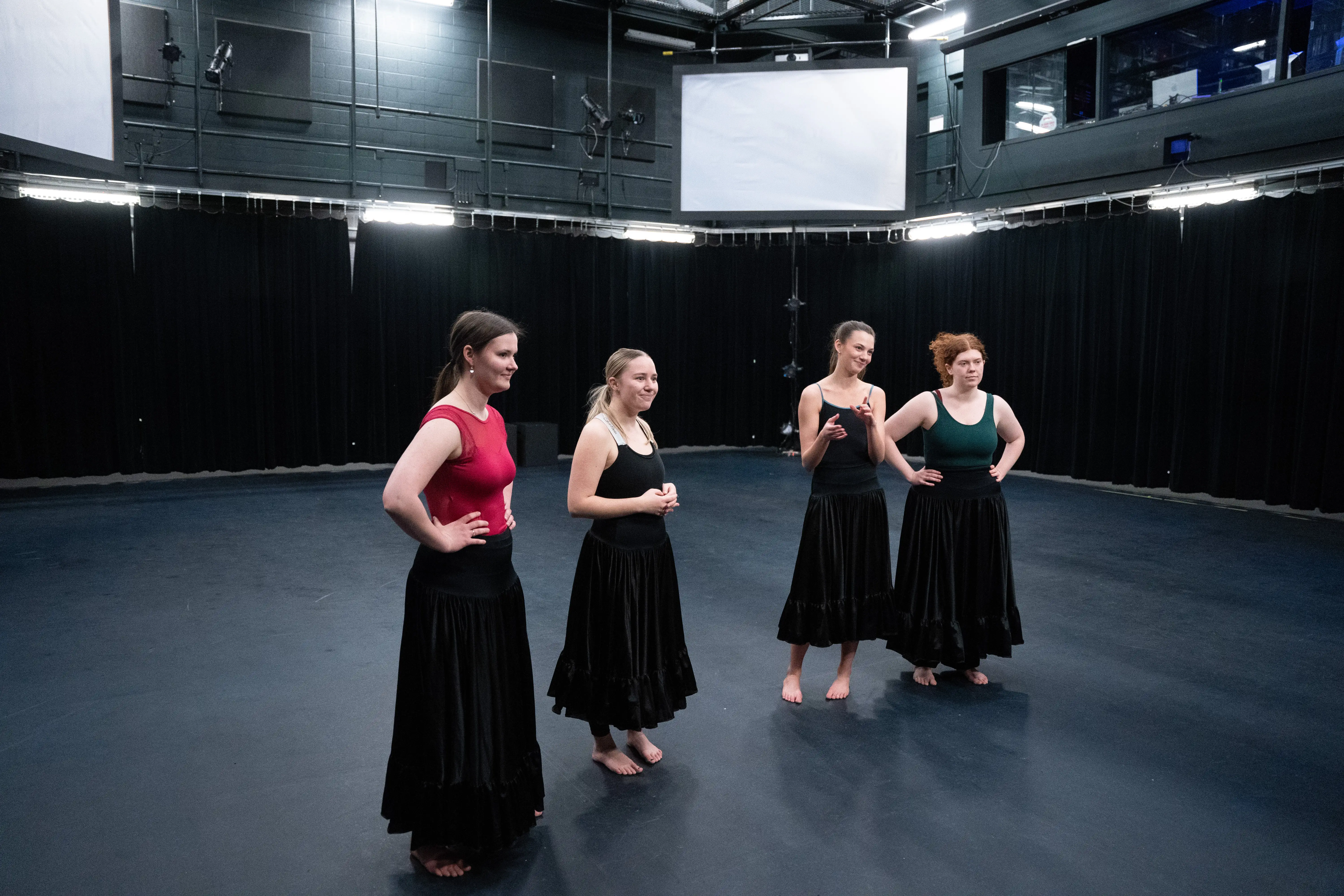 Four dance students in black skirts stand barefoot on stage in Belmont’s Black Box Theater, smiling and chatting after a rehearsal.