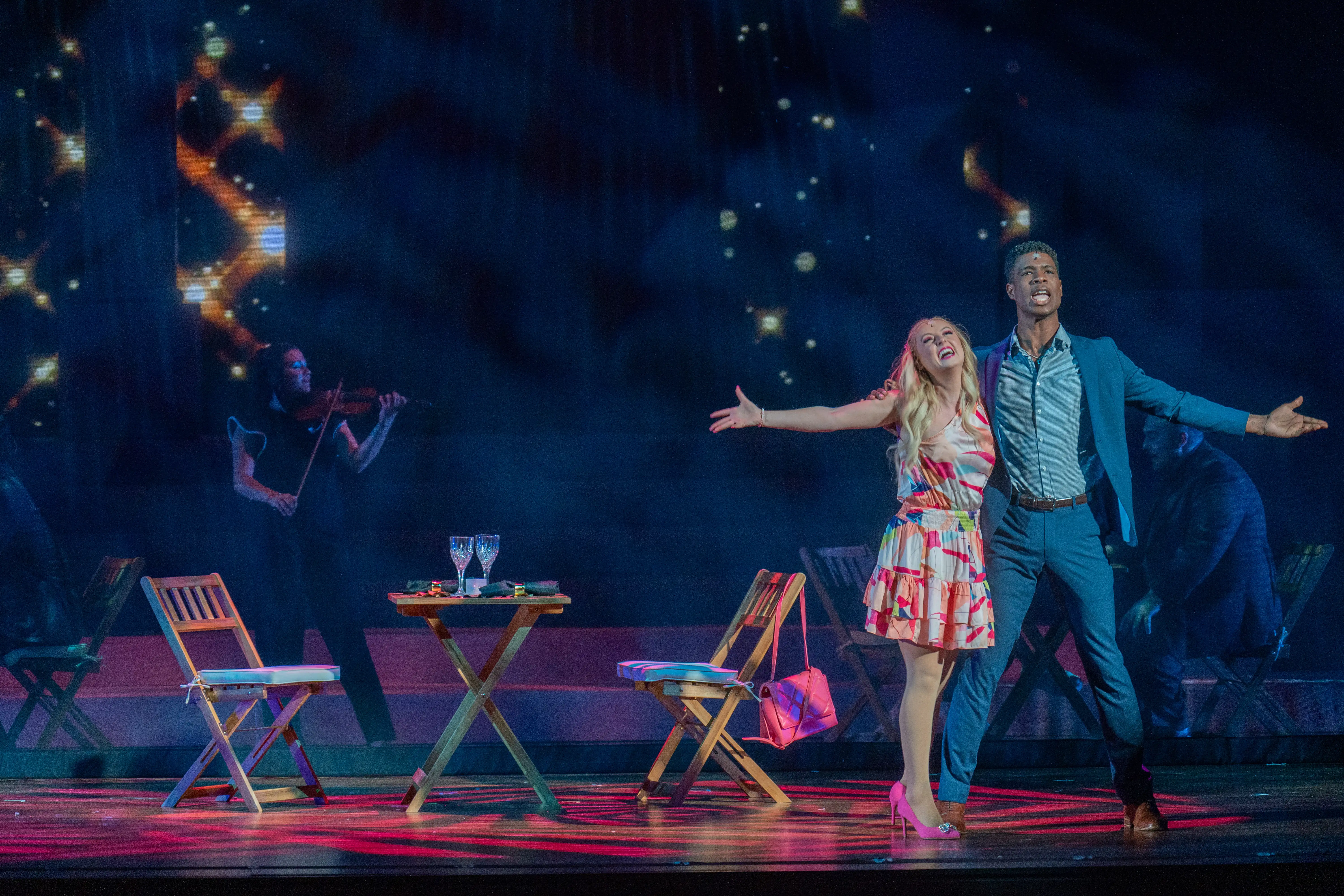 Two performers sing in a romantic duet at the Fisher Center, with a violinist silhouetted in the background and starry lights shining across the stage.