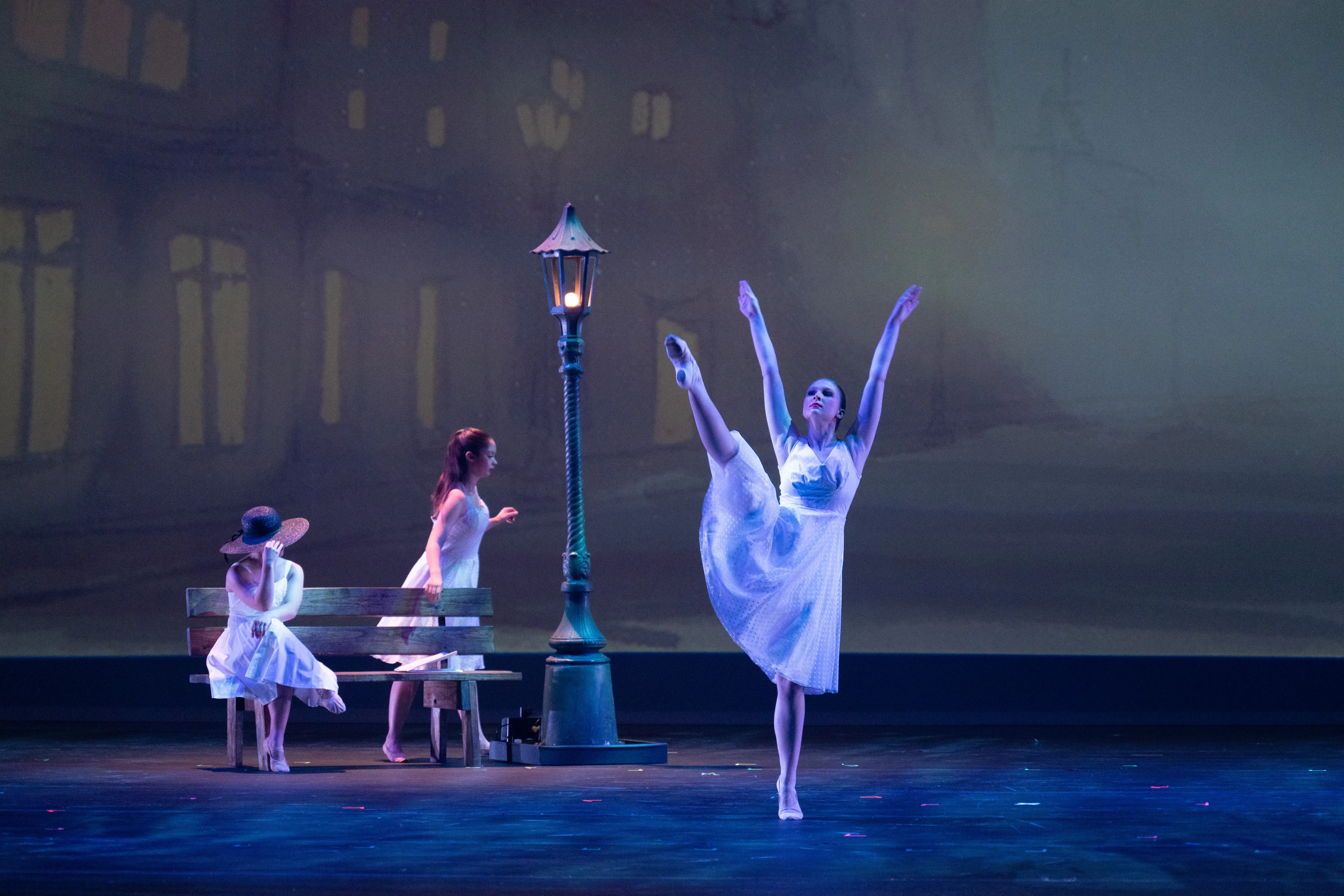 Ballet dancers in white dresses perform under soft blue lighting on the Fisher Center stage, beside a bench and vintage-style streetlamp.
