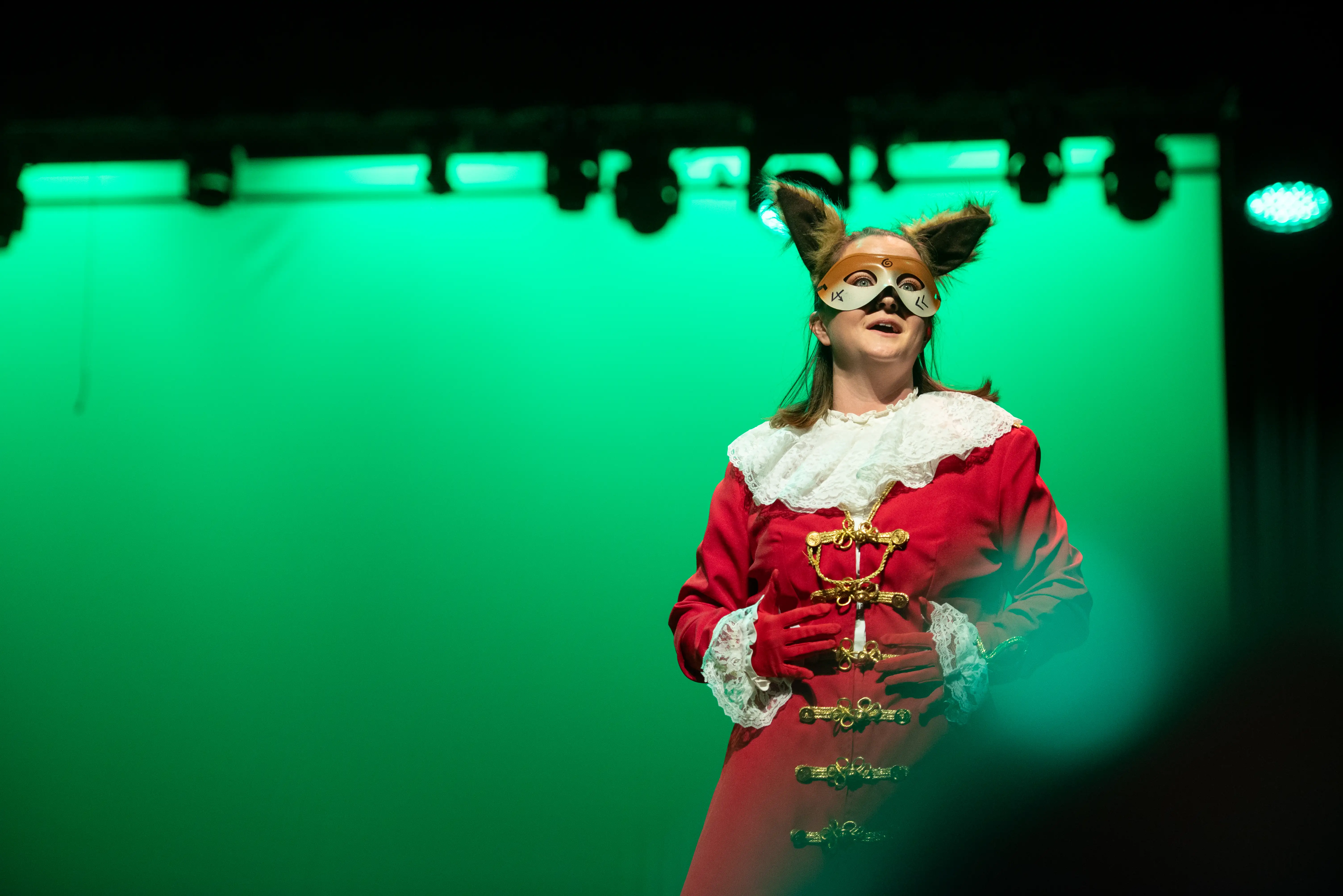 Performer in a red costume and fox mask stands under green stage lights at Massey Concert Hall, mid-speech during a dramatic production.