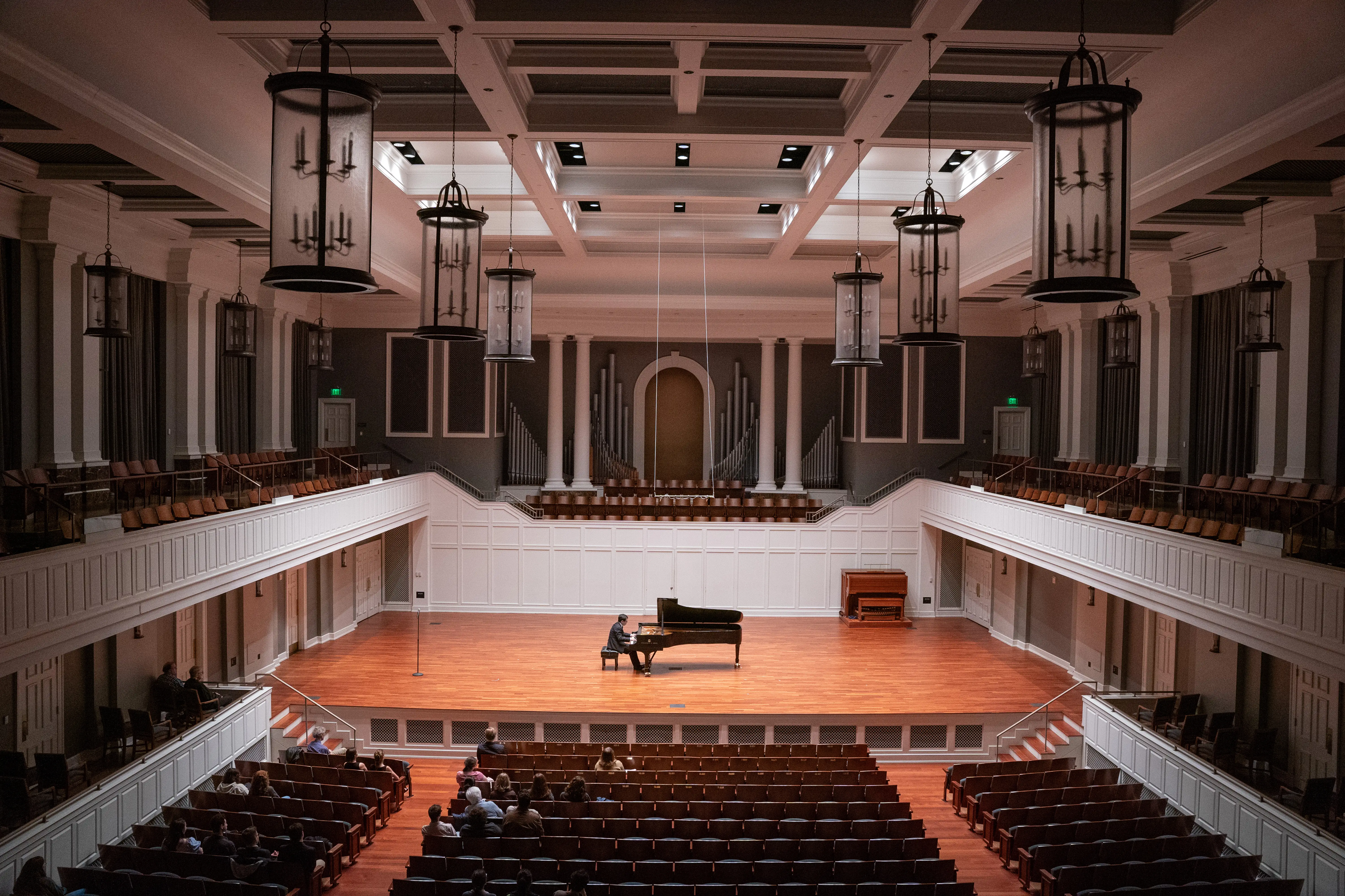 Audience watches a solo pianist perform onstage in McAfee Concert Hall, a bright, elegant venue with white walls and hanging lanterns.