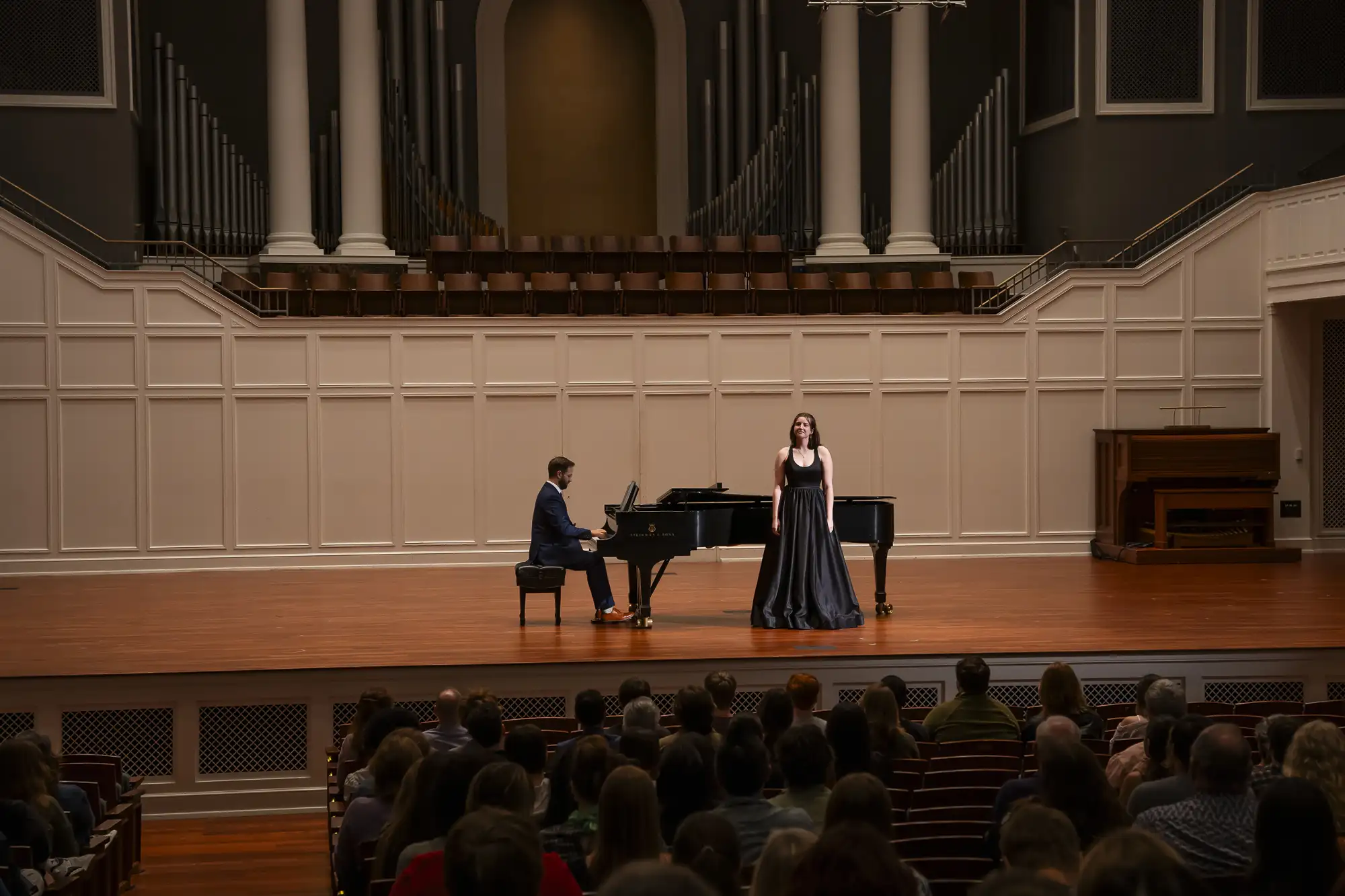 A vocalist performs with a pianist onstage in McAfee Concert Hall as an audience looks on, framed by the grand organ pipes and soft lighting.