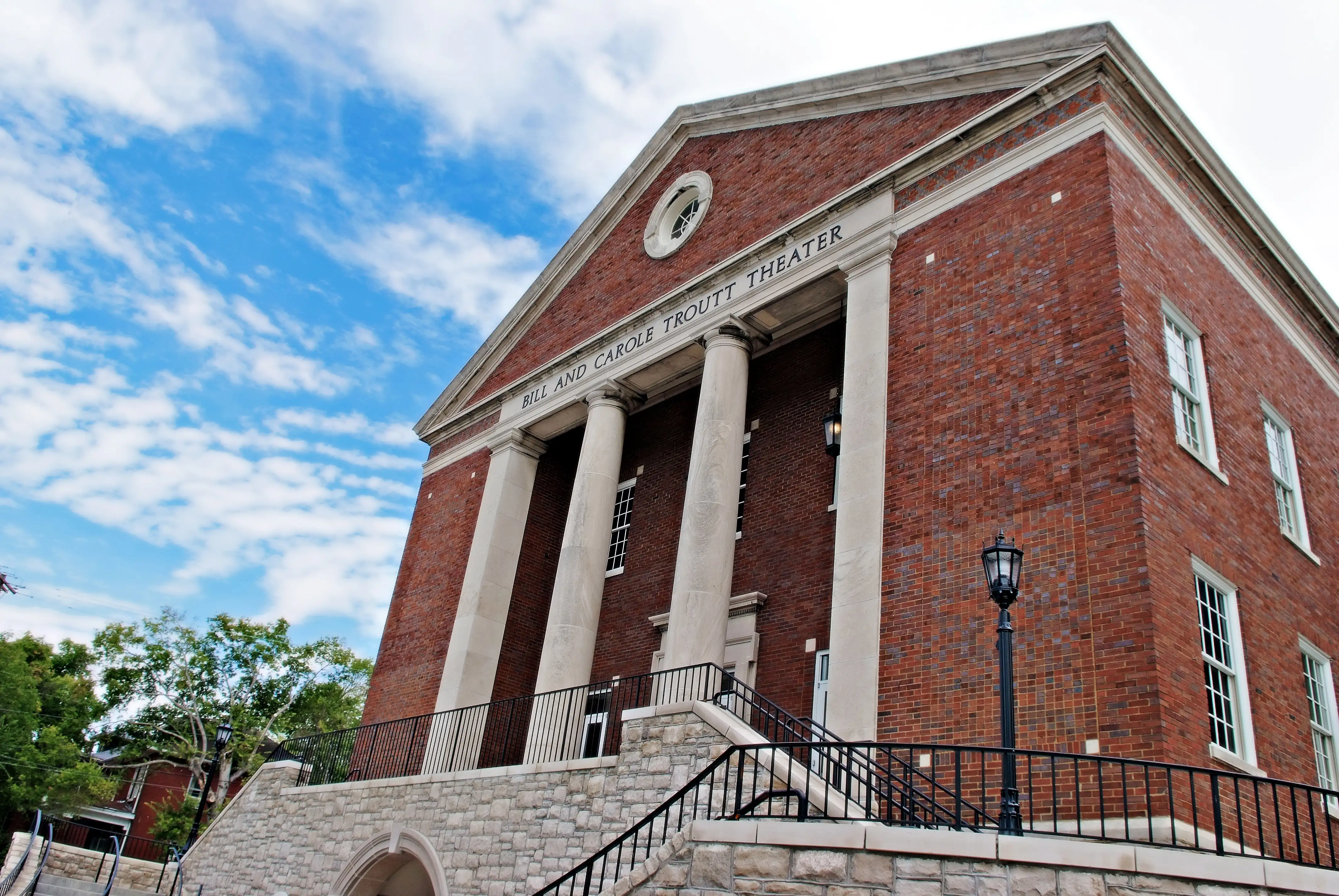 Exterior of Belmont’s Troutt Theater, a red brick building with white columns and stone steps under a partly cloudy sky.