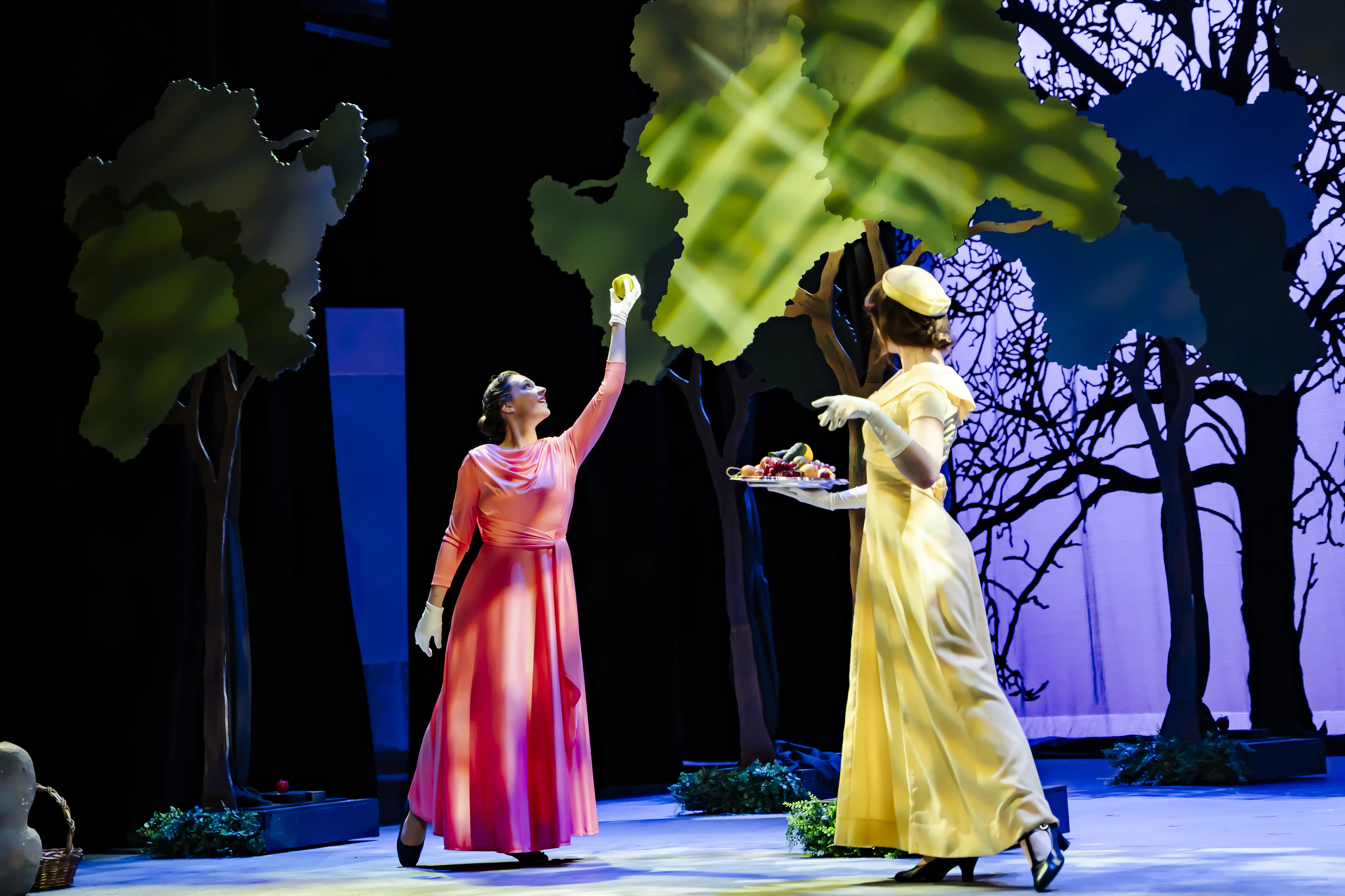 Two performers in bright dresses act out a garden scene with fruit and trees on a colorful set at Troutt Theater.