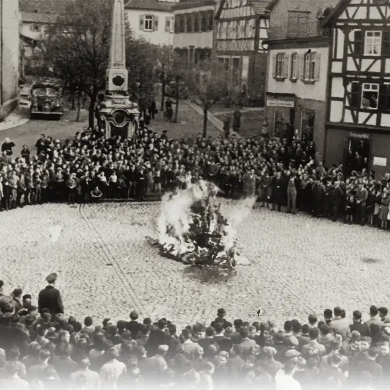 A historical black-and-white photograph showing a large crowd gathered around a bonfire in a cobblestone square. Buildings in the background are visible, along with trees. The scene depicts people observing the fire, some standing while others are seated, creating a sense of a communal event.