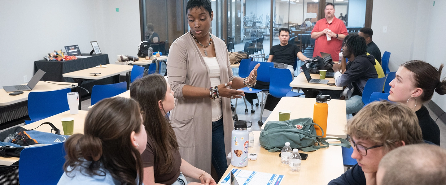 A female presenter speaking with students while they sit at a table with laptops during the Social Justice Hackathon.