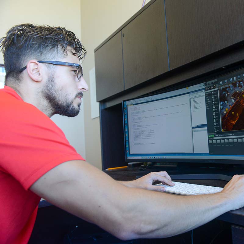 A young man with short, tousled hair and glasses is sitting at a desk, focused on a computer screen displaying code and graphics. He is wearing a red shirt and typing on a white keyboard, with an arm resting on the desk. The workspace appears well-lit, with a dark wooden cabinet in the background.