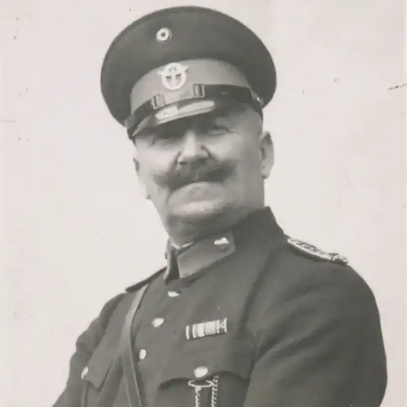  A black-and-white portrait of a smiling man in a police uniform, featuring a peaked cap and medals on his chest. He has a thick mustache and is posed slightly to the side, with a confident expression.