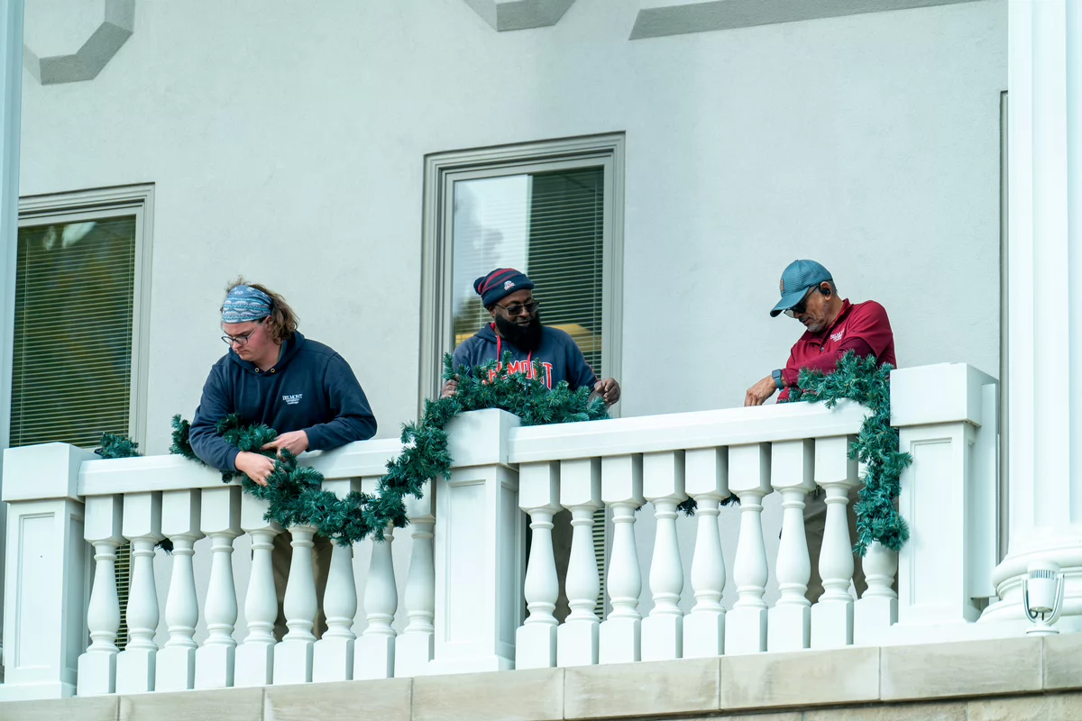 Three people decorate a balcony with holiday garlands. 