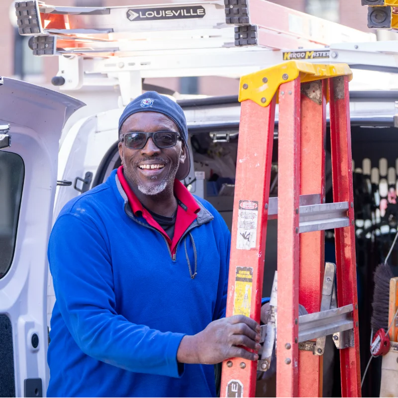 Smiling male technician in blue jacket, cap, and sunglasses, standing next to a red ladder with a service van.