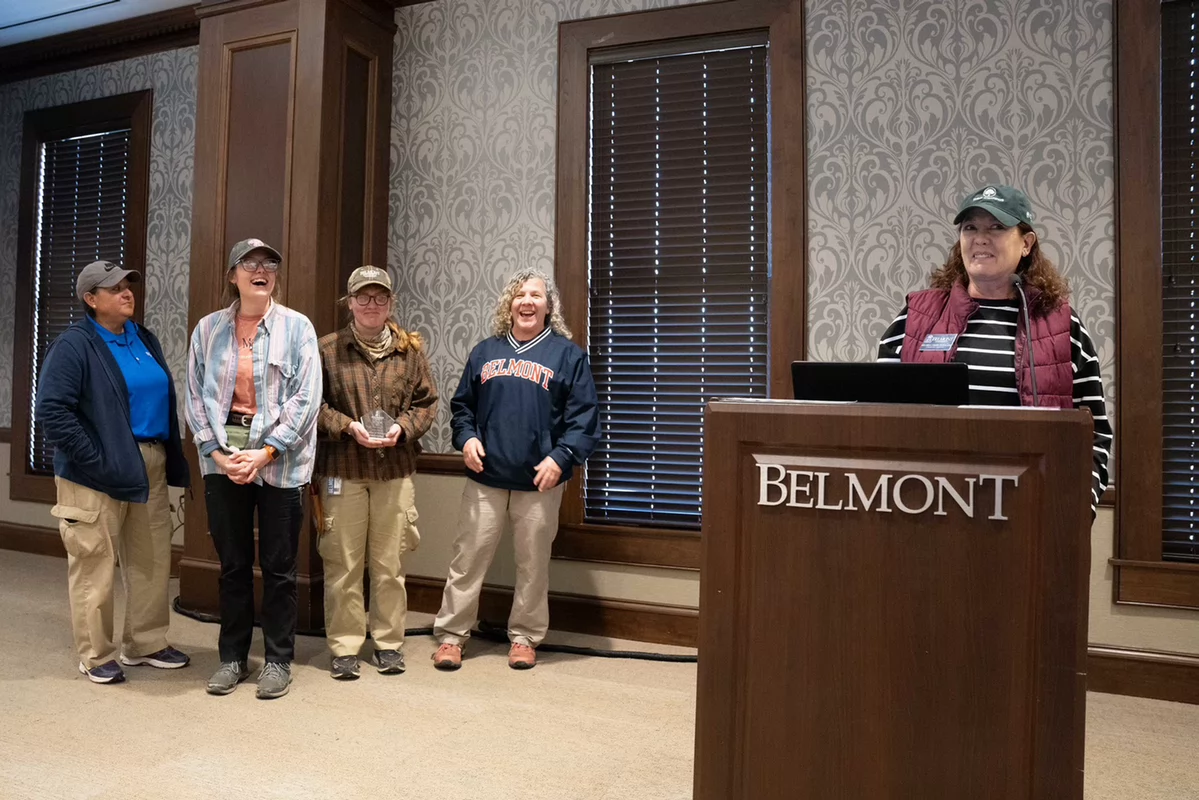 Five people are standing indoors at an event. Four are on the left, smiling, one holding a Team award. A woman stands at a podium on the right labeled "Belmont."