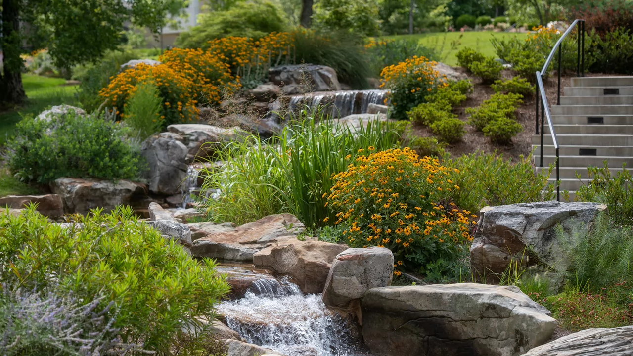 A serene garden scene with a small cascading waterfall flowing over rocks. Surrounding the stream are lush green plants and yellow flowers. Stairs with a railing ascend on the right.