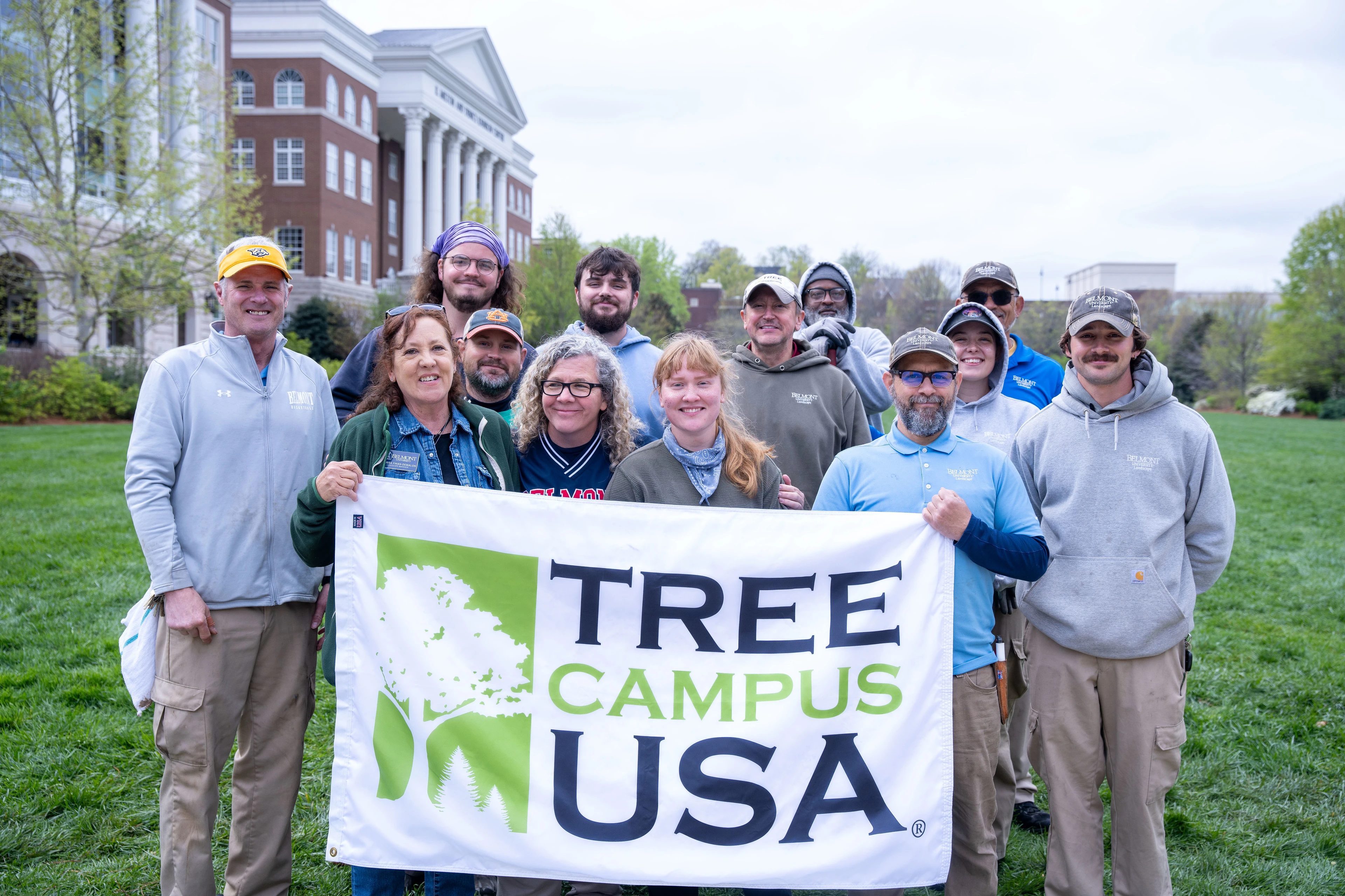 FMS team smiling and holding a "Tree Campus USA" banner outdoors. They stand on a lush green lawn with a large building in the background.
