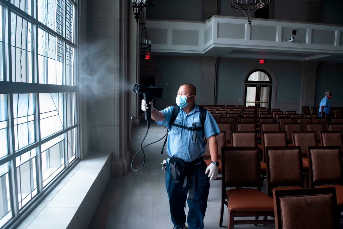 A person wearing a mask and gloves uses a fogger to disinfect an empty room with rows of chairs and large windows.