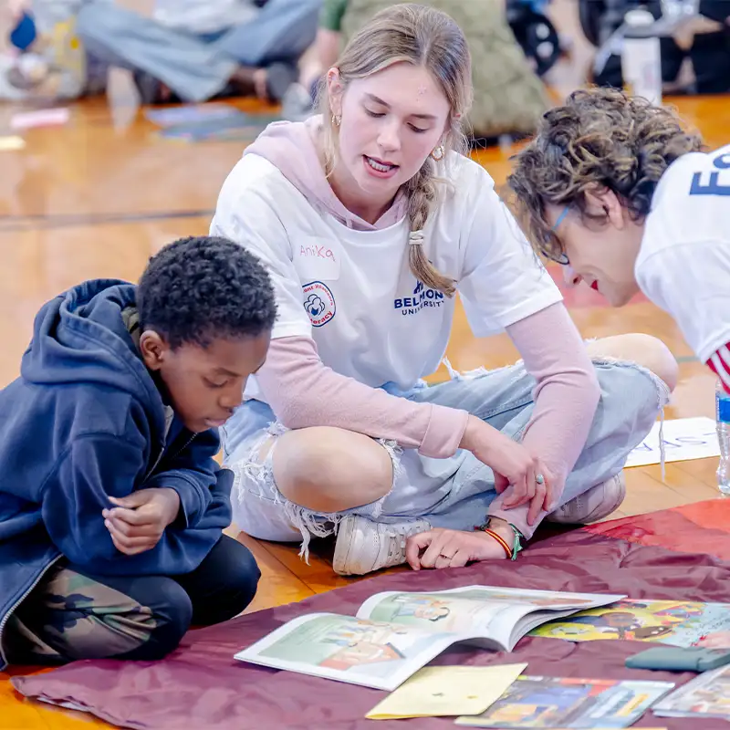 A Belmont student sitting cross legged with a couple of children who are also sitting on the floor reading books