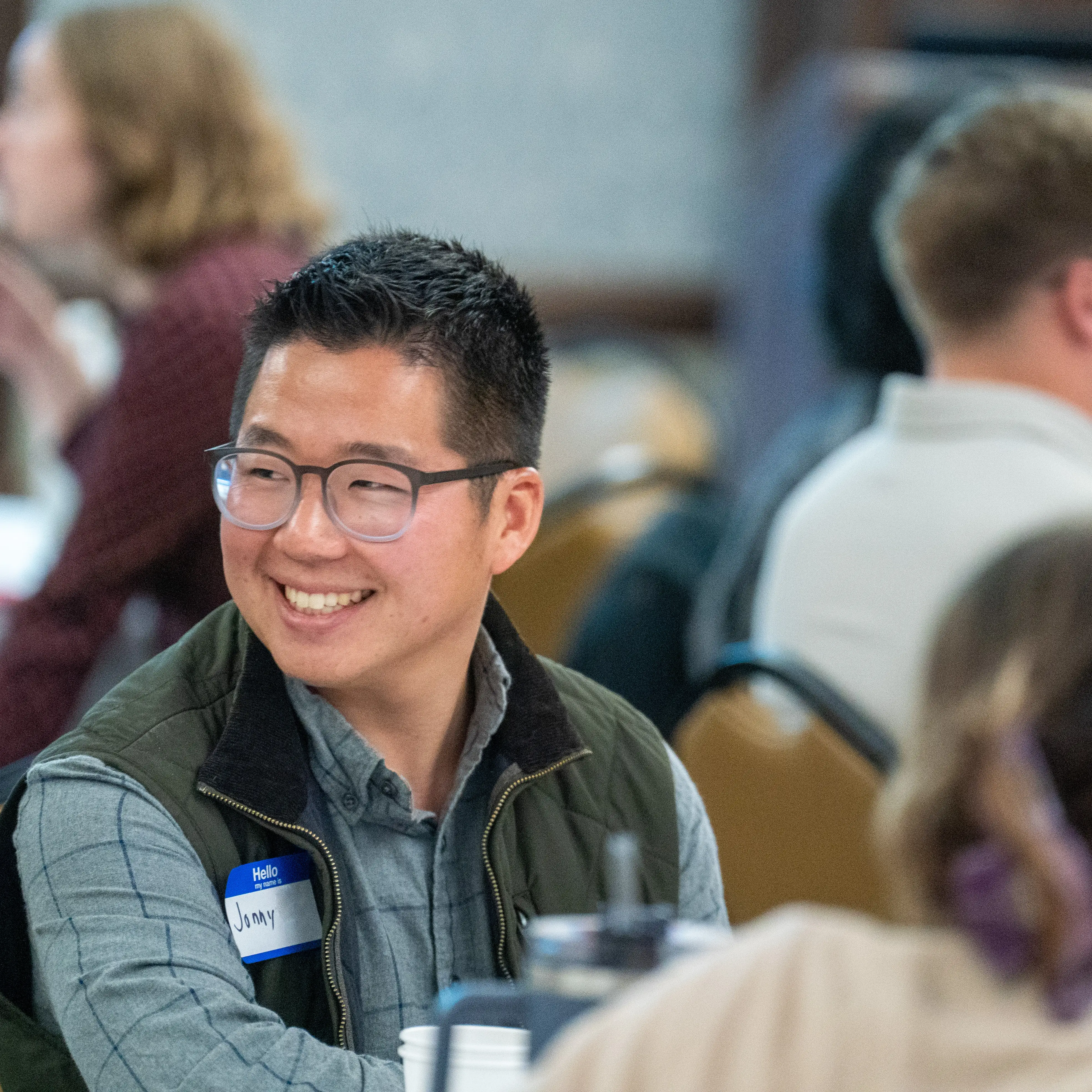 A student wearing a name tag smiles while talking with others at a table during a small-group gathering at Belmont University.