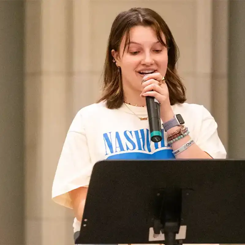 An indoor photo of a young woman holding a microphone and standing behind a music stand.