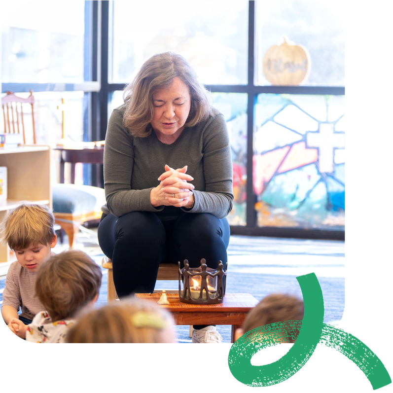 Rev. Susan Pendleton Jones leads a reflective moment with young children seated around her in a bright classroom. Sunlight streams through stained-glass windows in the background, creating a warm, peaceful atmosphere.