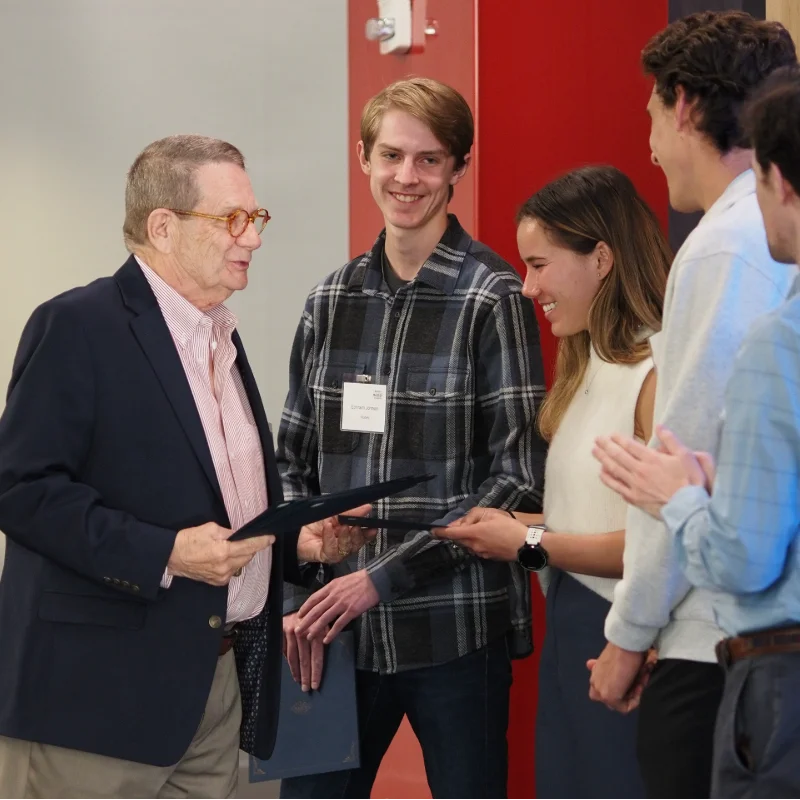 Man presenting a certificate to a smiling student, while others applaud at an academic recognition ceremony.