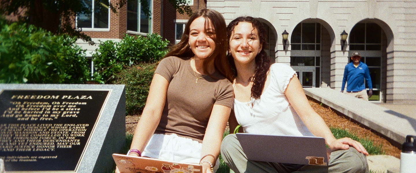 Two smiling students sit outside on a sunny day in front of a building with arched windows, using laptops near a plaque labeled “Freedom Plaza.” A person walks in the background.