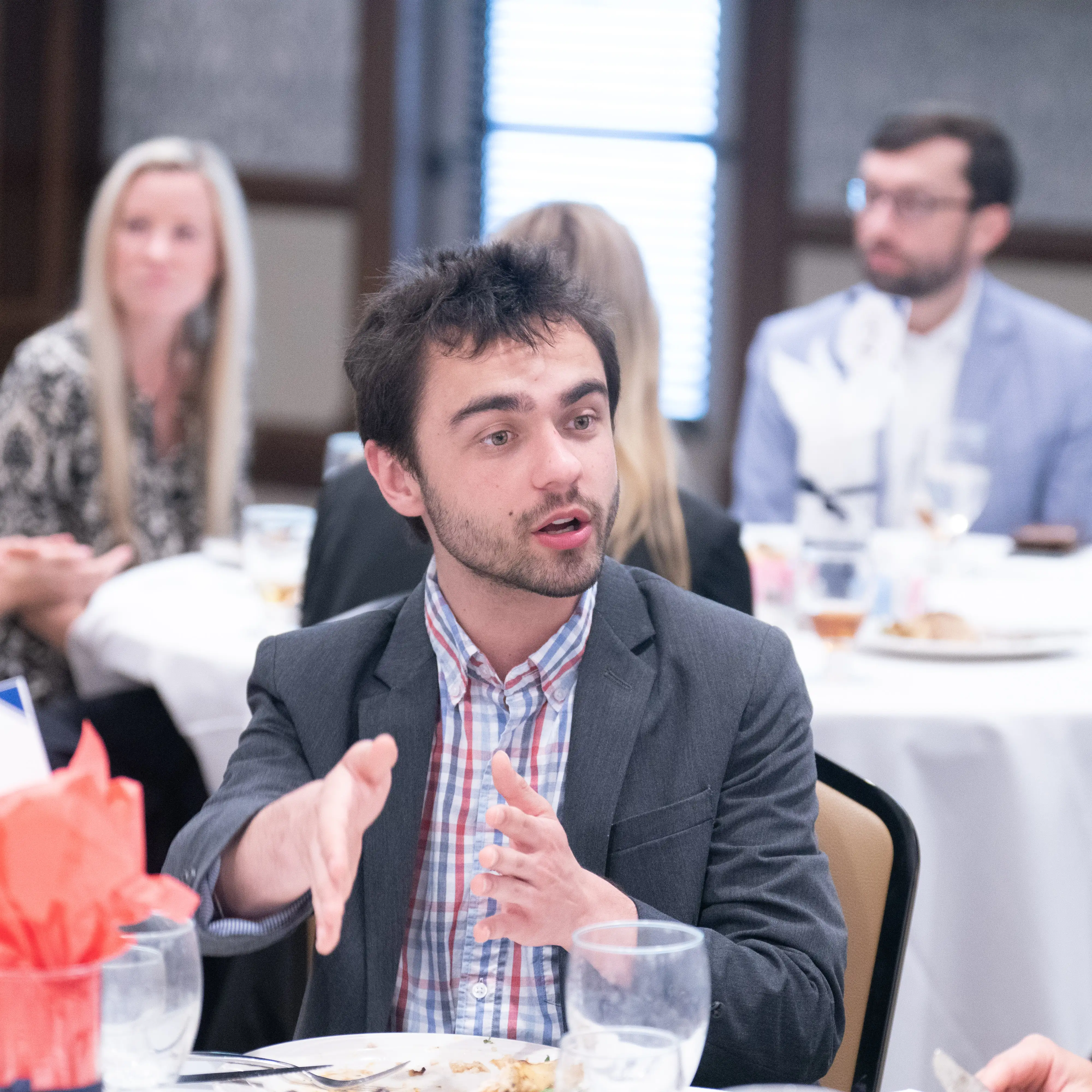 A Belmont student speaks while seated at a round table during a small group discussion or luncheon, gesturing as others listen in the background.
