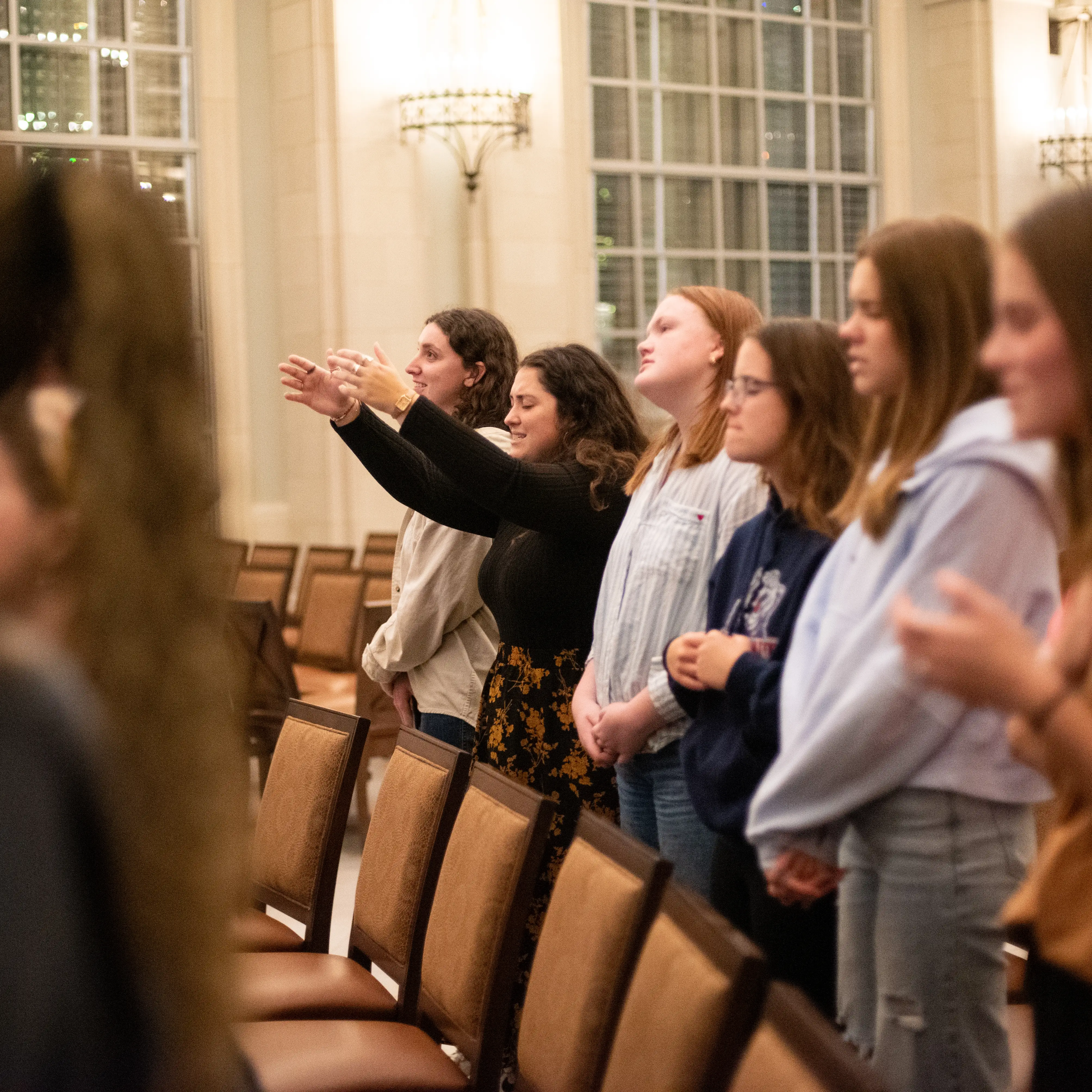 Students stand in rows during a worship service, some with hands raised, inside a softly lit chapel on Belmont University’s campus.