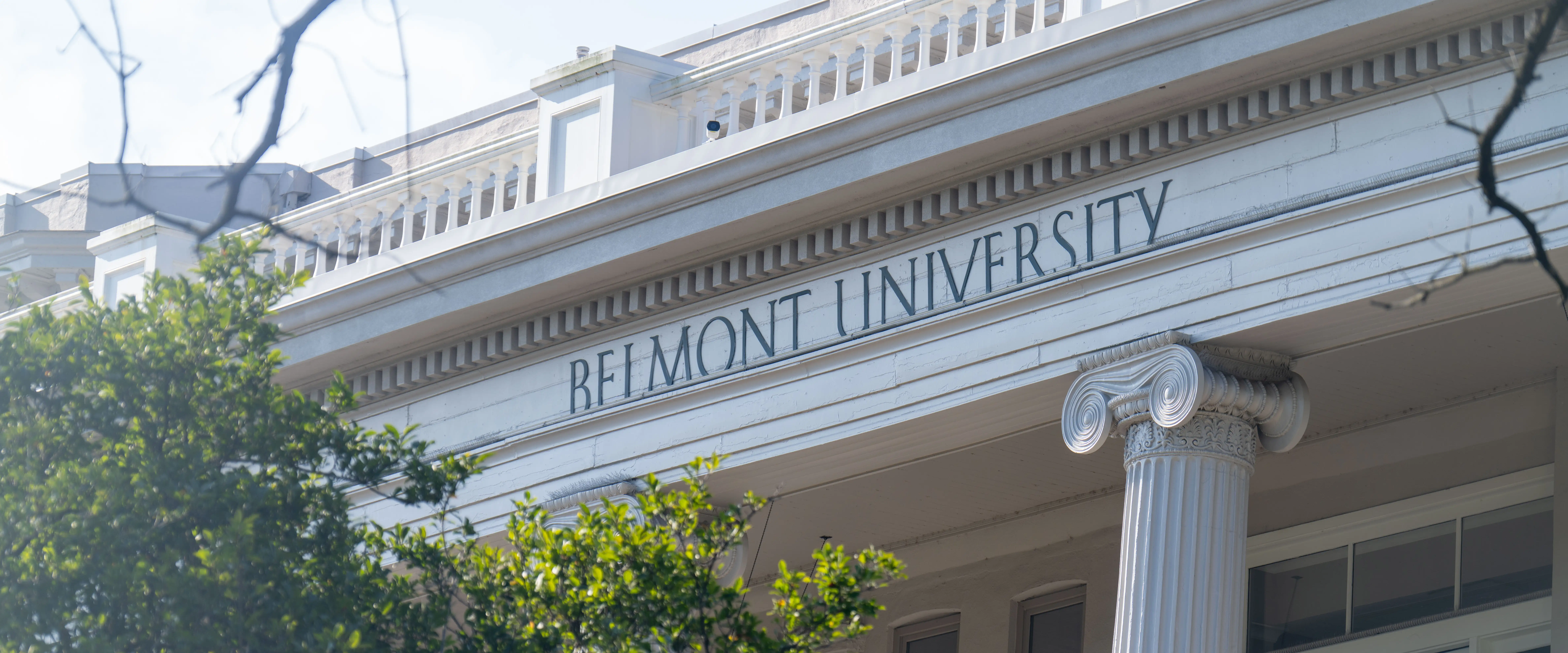 The Belmont University name is carved into the pediment of a white, columned campus building, partially framed by green trees on a sunny day.