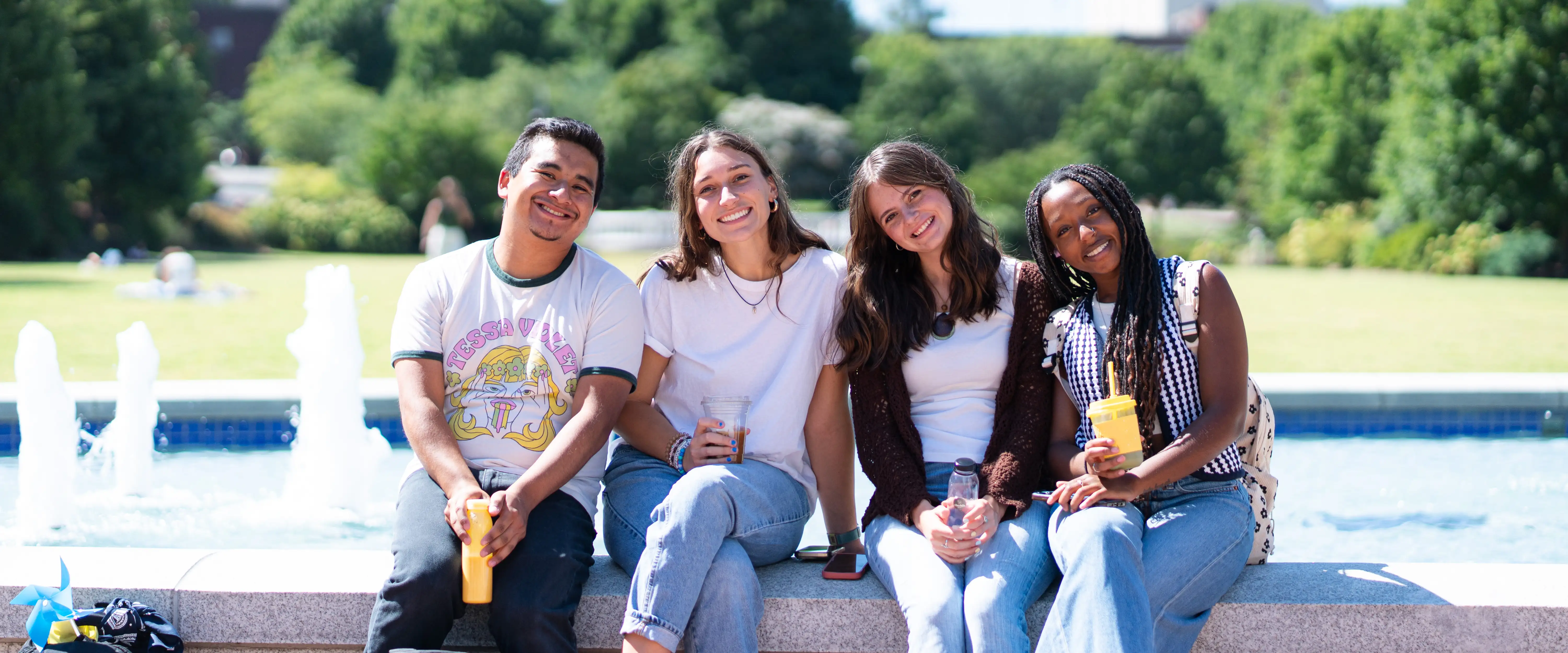 Four students sit together on a low stone wall near a campus fountain, smiling and holding drinks on a sunny day at Belmont University.