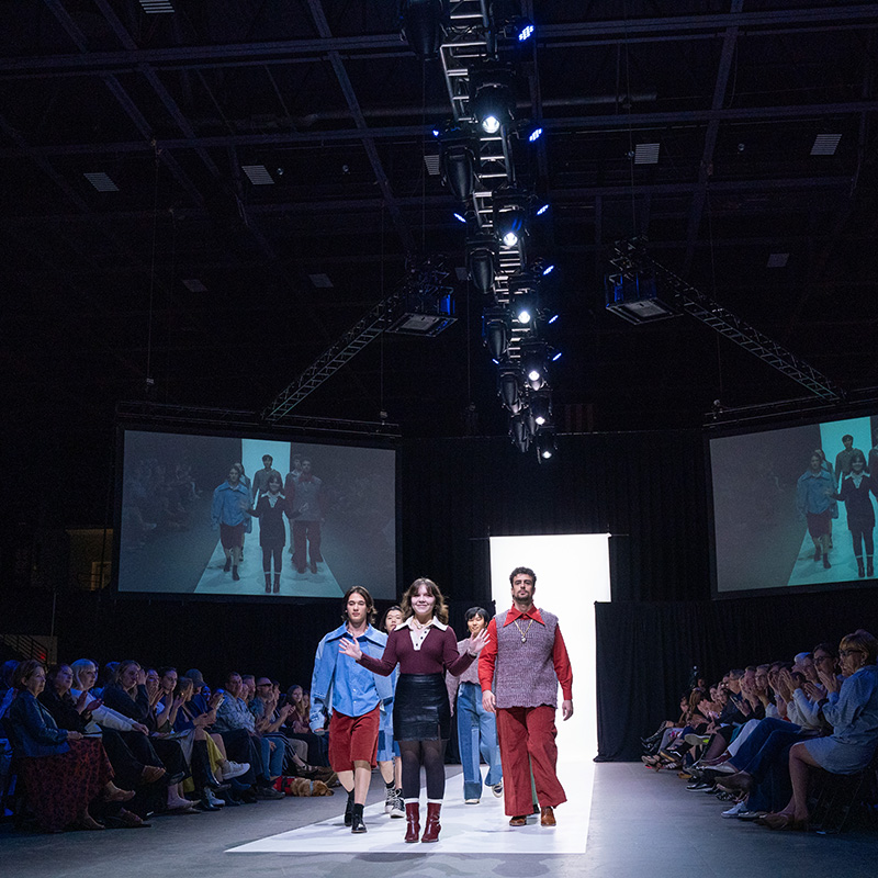 Models walk down a brightly lit runway at a fashion show, with the student designer waving to the audience. The venue is dimly lit except for the stage lights overhead, which highlight the models and their outfits. Two large screens on either side of the runway display a live feed of the show. The audience, seated along both sides of the runway, watches and applauds.