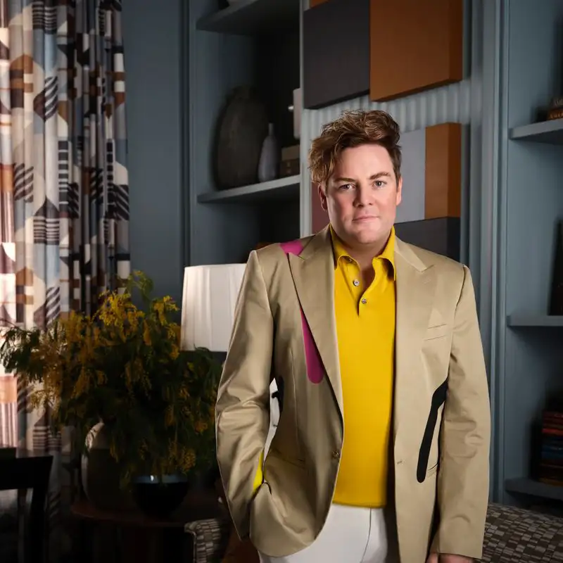 Jonathan Savage wearing a tan blazer with colorful accents and a yellow shirt stands in a stylish room with patterned curtains, shelving, and a floral arrangement.
