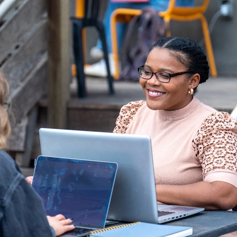 Smiling woman with glasses working on a laptop at an outdoor table.
