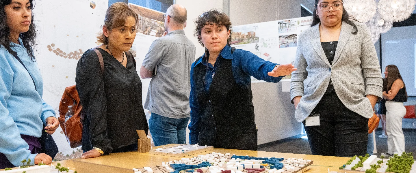 A group of architecture students or designers reviewing a detailed urban planning model, with one person presenting.