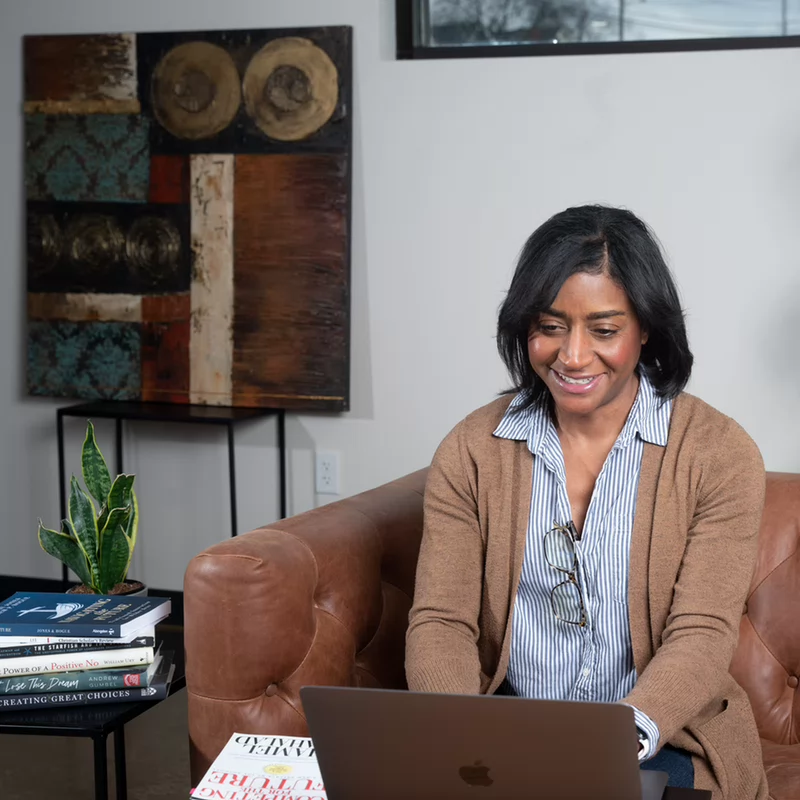 A person sits casually on a stone bench near a large window, focused on a laptop. Backpacks are beside them, suggesting a study or work session.