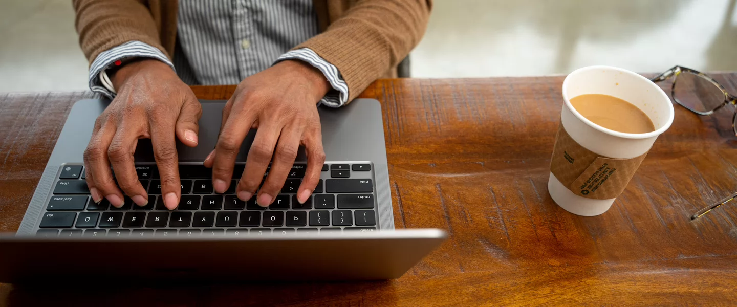 A person in glasses works intently on a laptop at a café table. Colorful pens and a tumbler are nearby. The atmosphere is focused and busy.