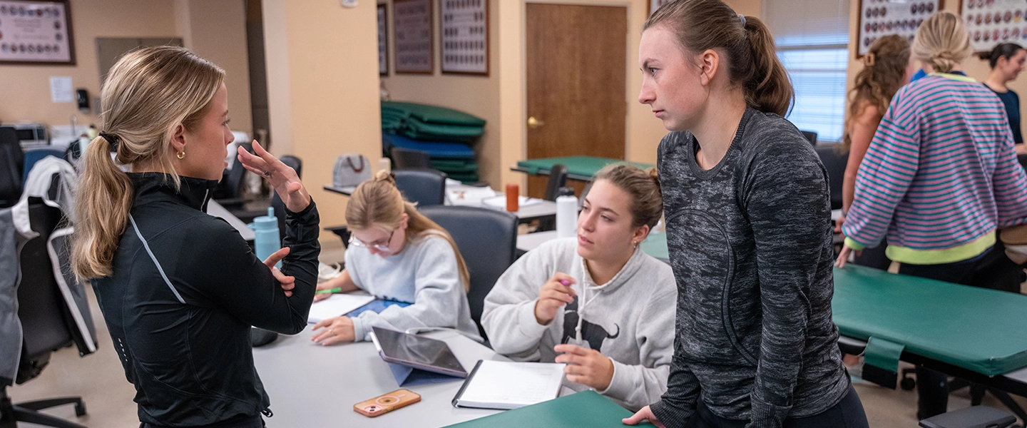 Students collaborate in a clinical classroom, discussing notes at a table while treatment tables and medical equipment are visible in the background.