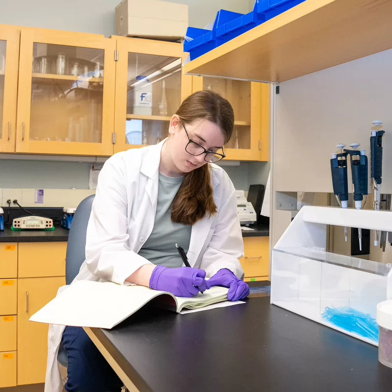 A woman in a lab coat and purple gloves writes in a notebook in a laboratory. She is surrounded by shelves with lab equipment, conveying focus and concentration.