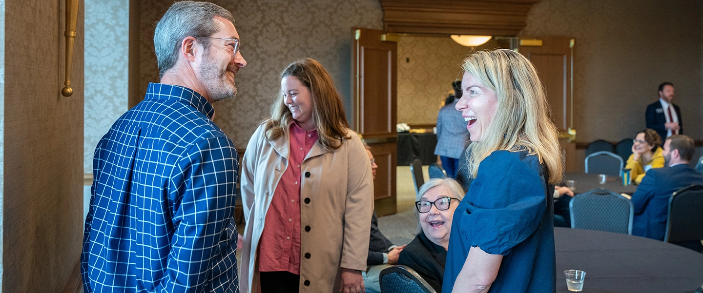A group of faculty smiling and chatting around a table in a warmly lit conference room. One man in a blue plaid shirt faces three women, including one seated, all engaged in conversation. In the background, others are gathered near tables and chairs.