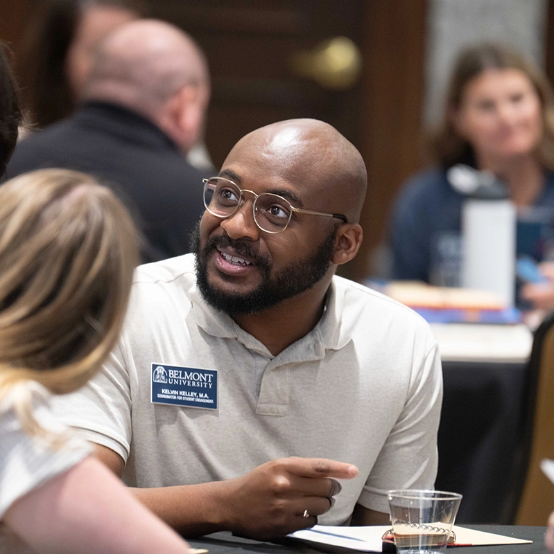 A Belmont University staff member wearing glasses and a name tag  smiles and engages in conversation during a group meeting.