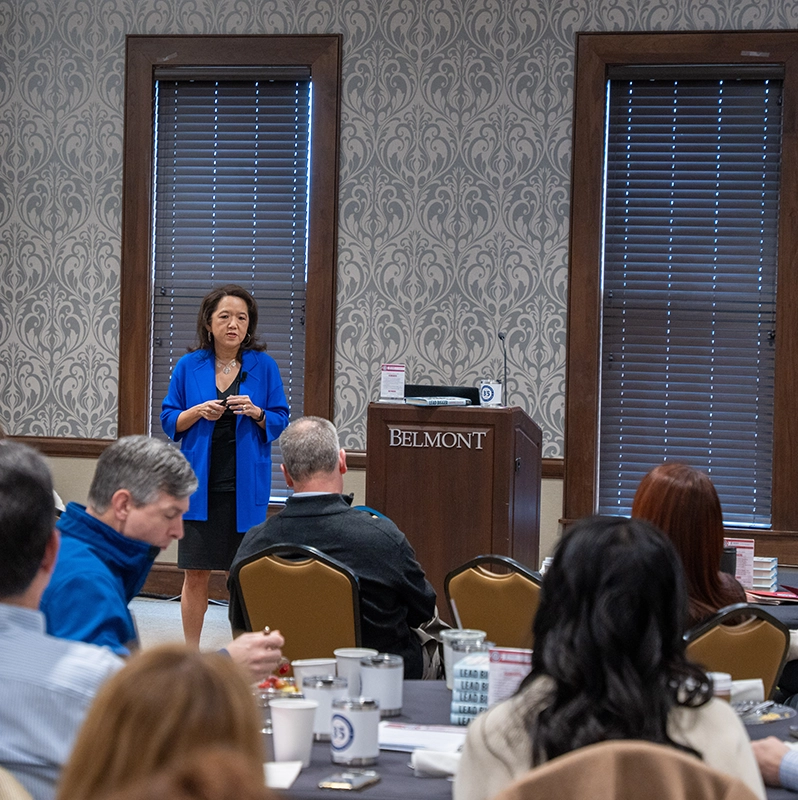 A woman in a blue blazer speaks to a seated audience during a presentation at Belmont University, standing next to a podium with the Belmont logo.