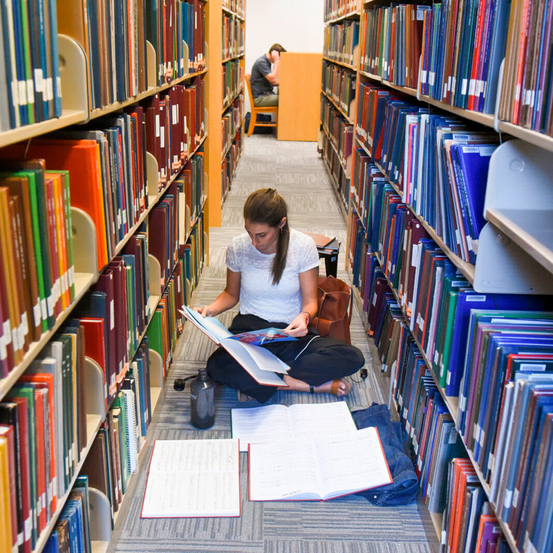 A student seated on the library floor between two bookshelves.