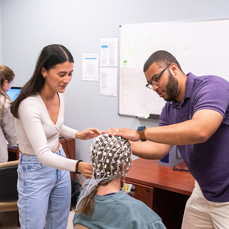 Belmont University students conducting a neuroscience experiment, fitting an EEG cap on a participant in a research lab.