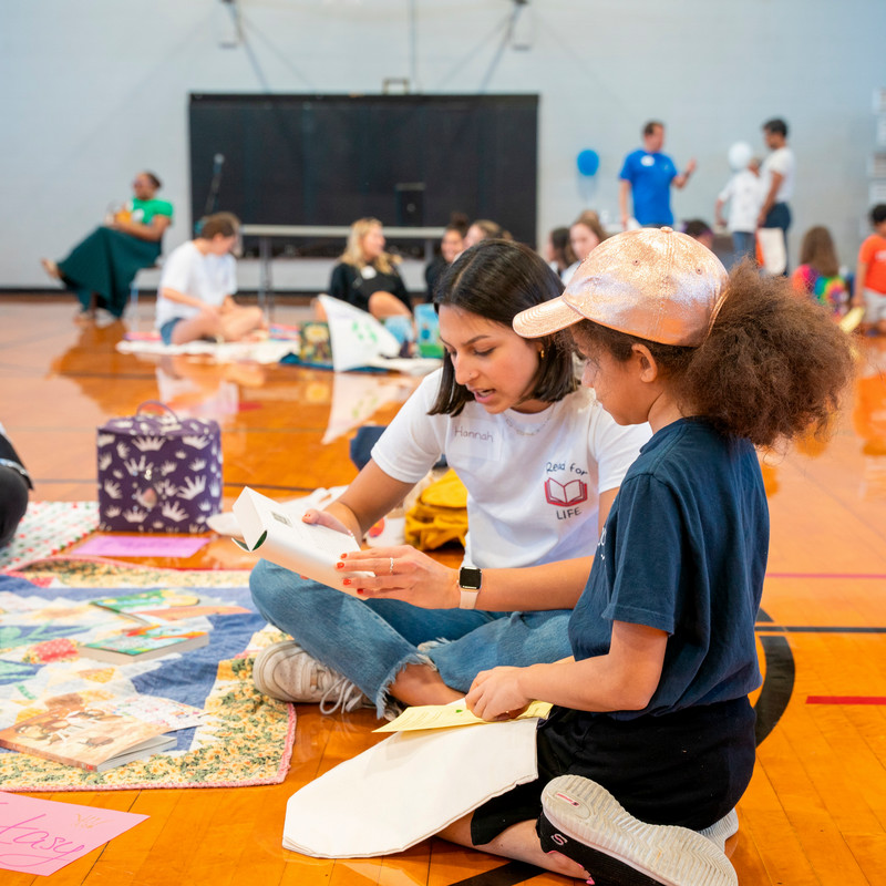 student sitting on the floor of a gym reading to a young child